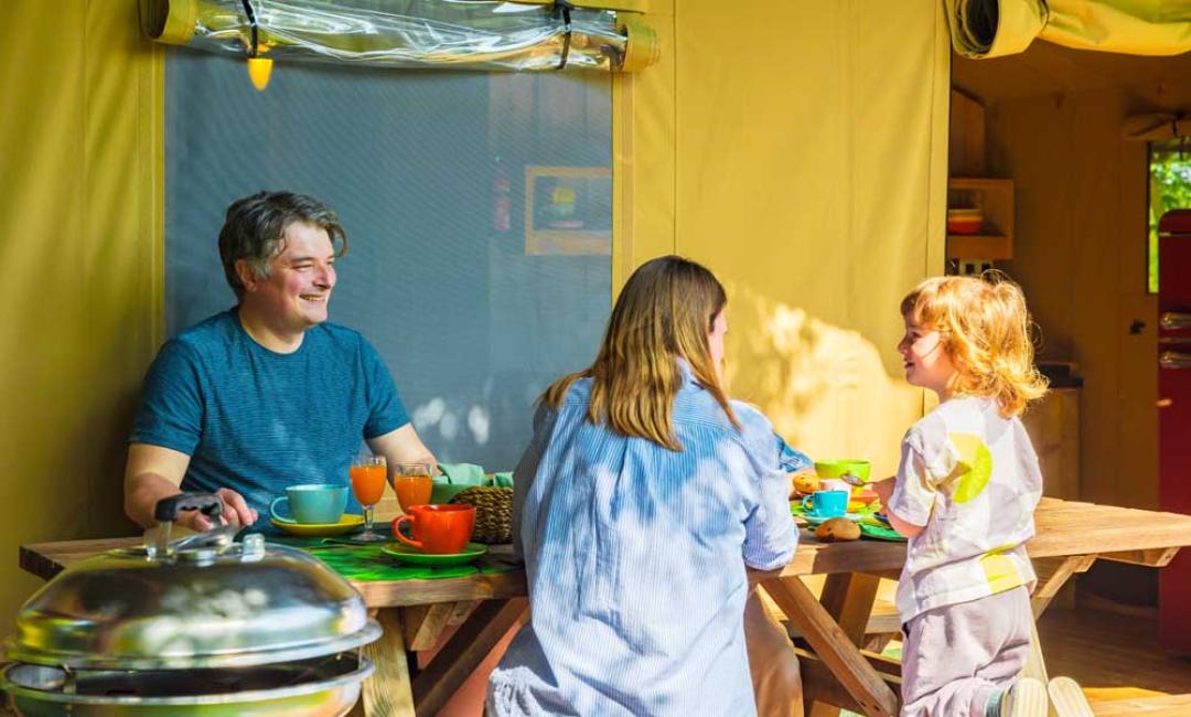 Familie spiser morgenmad på campingplads, smiler og nyder tiden sammen.